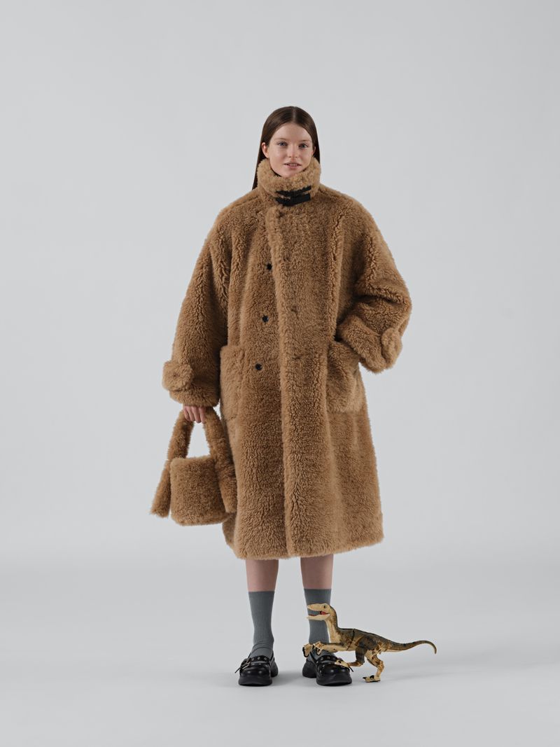 A young woman poses in a full-length shot, wearing an oversized brown faux-fur coat with a matching purse, against a neutral background.
