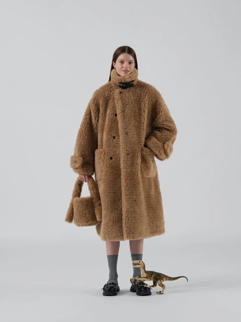 A young woman poses in a full-length shot, wearing an oversized brown faux-fur coat with a matching purse, against a neutral background.