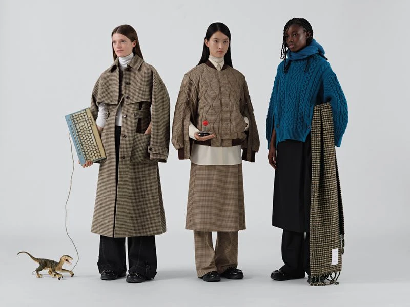 Three fashion models in studio, wearing patterned coats and skirts, standing next to a toy dinosaur, computer keyboard, and scarf.