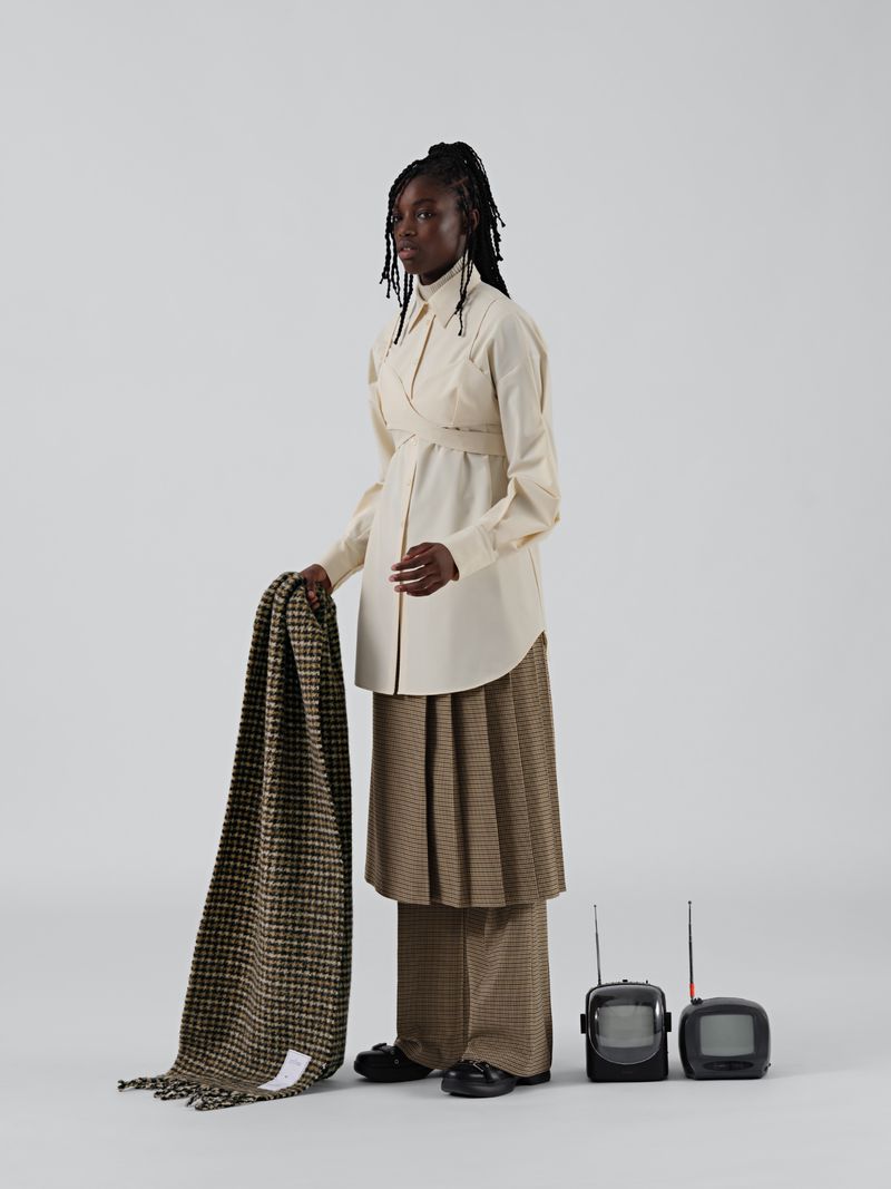 An African woman in a stylish cream wrap shirt, wide plaid pants, and black loafers holding a brown blanket with two small retro TVs.