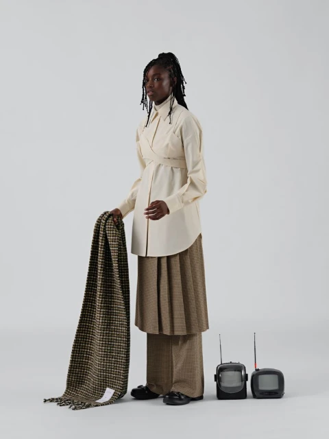 An African woman in a stylish cream wrap shirt, wide plaid pants, and black loafers holding a brown blanket with two small retro TVs.