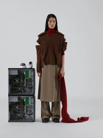 Fashion shot of a slender Asian woman in a brown sweater, tan skirt, and a red scarf, standing next to stacked computer towers in a studio.