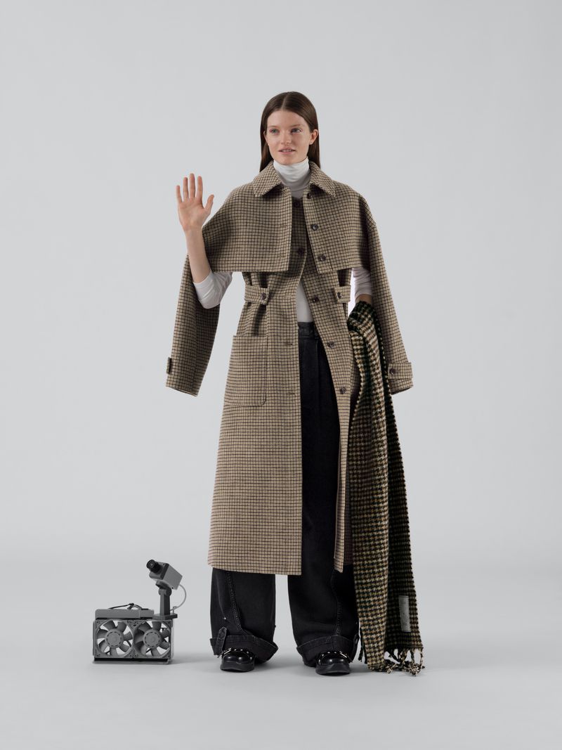 Model wearing a plaid trench coat and dark trousers waving against a seamless light grey background with a small portable fan on the floor.