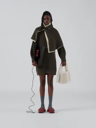 Model wearing a layered olive green knit sweater with cream trim and holding a white fluffy handbag against grey background
