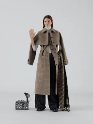 Model wearing a plaid trench coat and dark trousers waving against a seamless light grey background with a small portable fan on the floor.