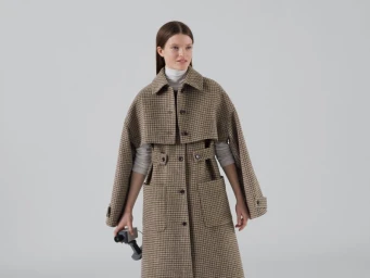 Model wearing a plaid trench coat over a white turtleneck holding a camera against a white seamless background