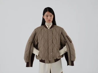 Model wearing a brown quilted bomber jacket with arm zippers over a cream shirt against a white seamless background