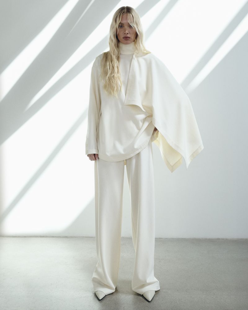 A fair-skinned, blonde adult woman stands in a light studio wearing matching cream-colored, high-necked top, palazzo pants, and pointed heels.