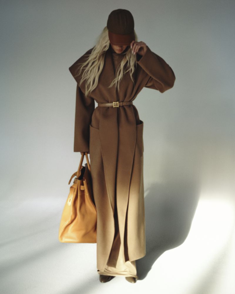 Model Elsa Hosk wearing a long brown belted coat and cap, holding a large tan leather handbag against a white studio background.