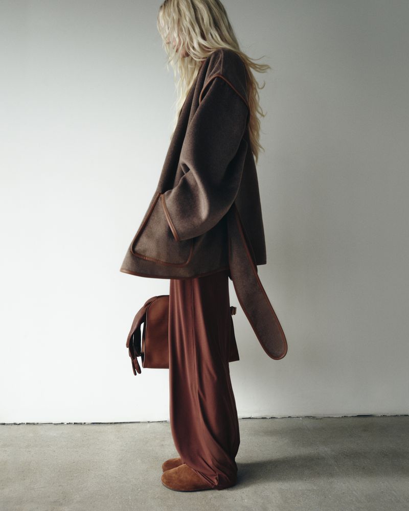 Model Elsa Hosk wearing a brown coat and trousers holding a bag against a white seamless background