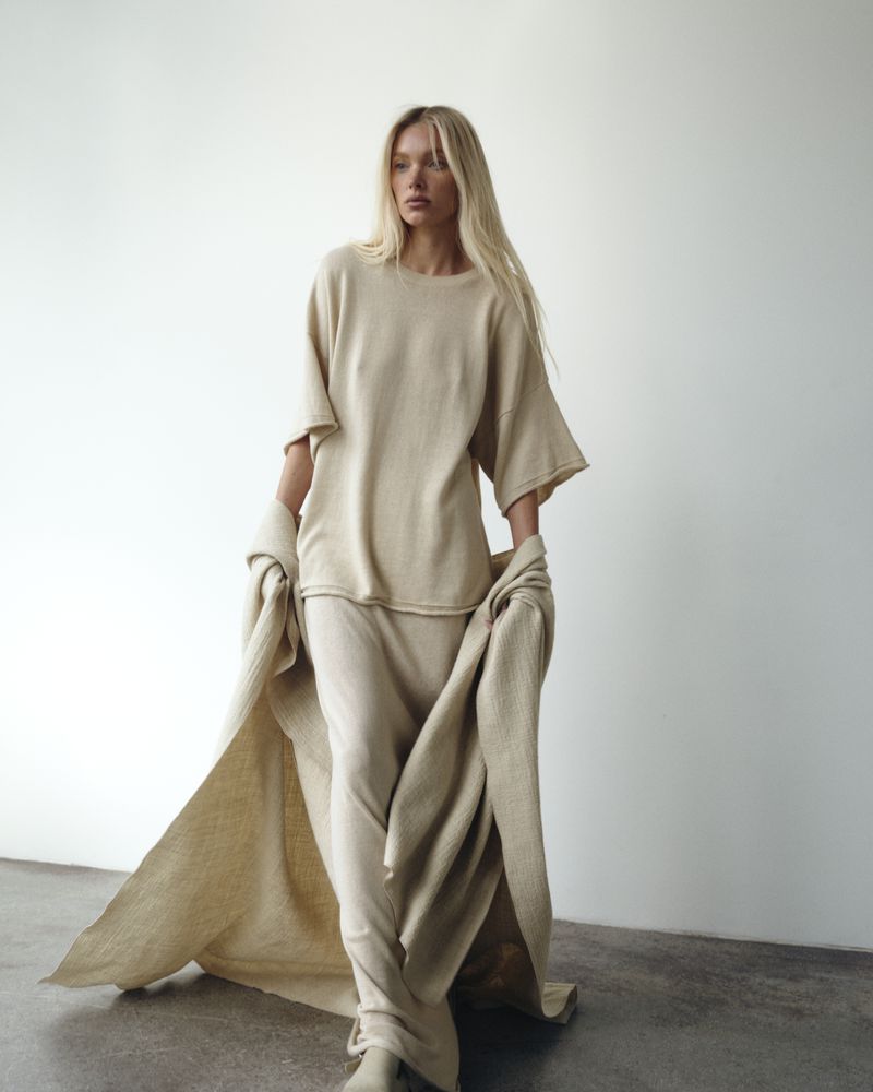 Model Elsa Hosk wearing a tonal cream knit outfit and draped throw against a plain white studio wall