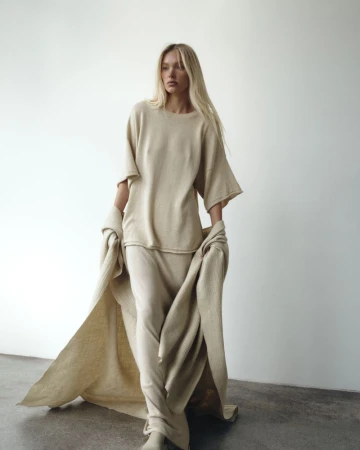 Model Elsa Hosk wearing a tonal cream knit outfit and draped throw against a plain white studio wall