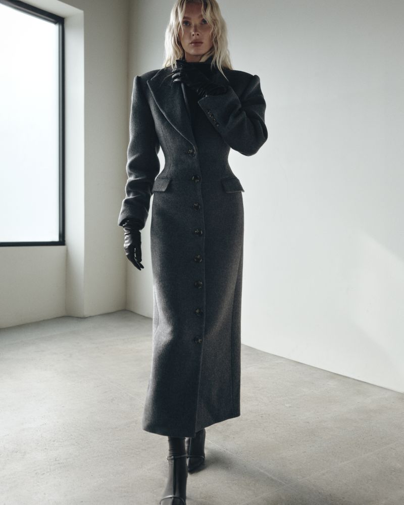 Elegant woman in a long grey button-down coat, black gloves, and heeled boots walking indoors with neutral walls and a large window.