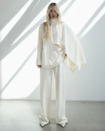 Model Elsa Hosk wearing a white turtleneck and trousers against a wall with strong diagonal shadows.