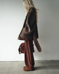 Model Elsa Hosk wearing a brown coat and trousers holding a bag against a white seamless background