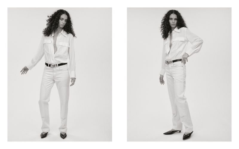 Model with long dark curly hair wearing a white button-down shirt and white pants against a seamless light background.