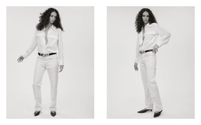 Model with long dark curly hair wearing a white button-down shirt and white pants against a seamless light background.