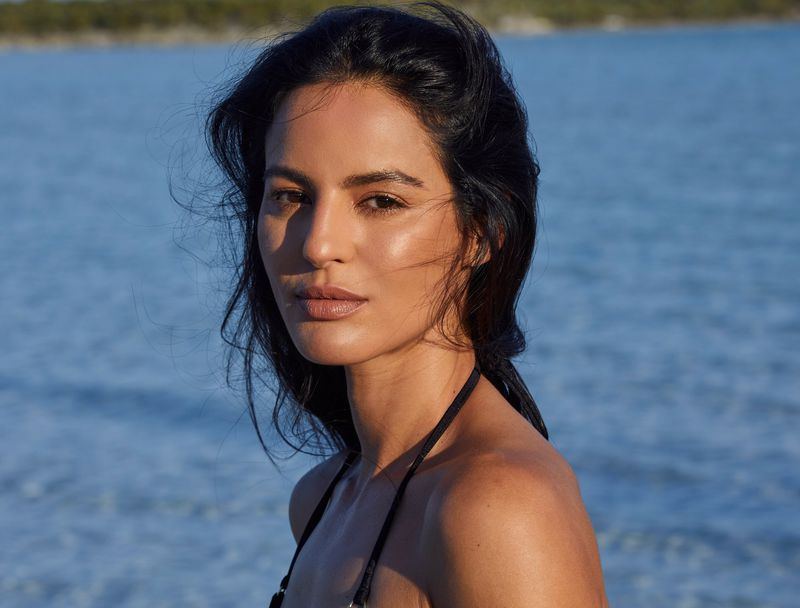 Close-up of a woman with dark hair posing against a blurred blue water background, creating a portrait with natural light and clear focus.