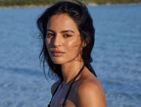 Close-up of a woman with dark hair posing against a blurred blue water background, creating a portrait with natural light and clear focus.