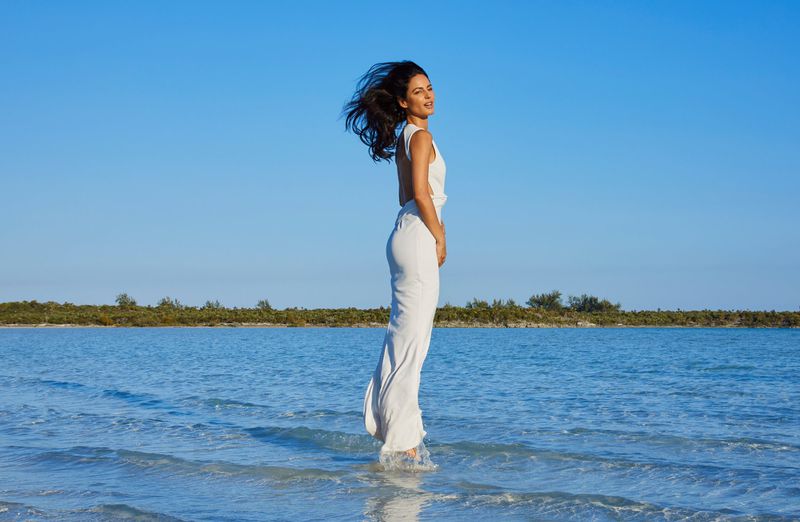 Adult woman in a white dress stands in shallow ocean water with clear blue sky in an outdoor setting under bright sunlight.