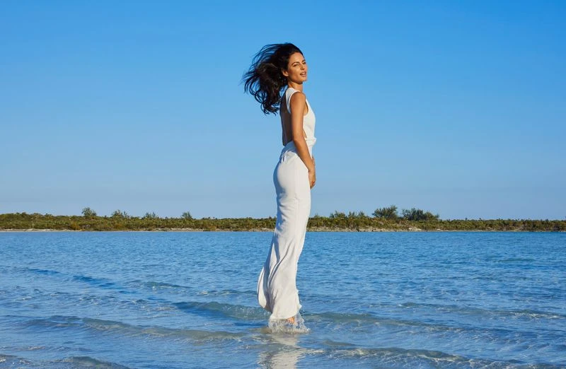 Adult woman in a white dress stands in shallow ocean water with clear blue sky in an outdoor setting under bright sunlight.
