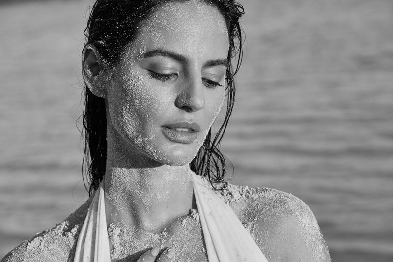 Black and white portrait of a woman with closed eyes, her face and shoulders covered in sand at the beach near the ocean.