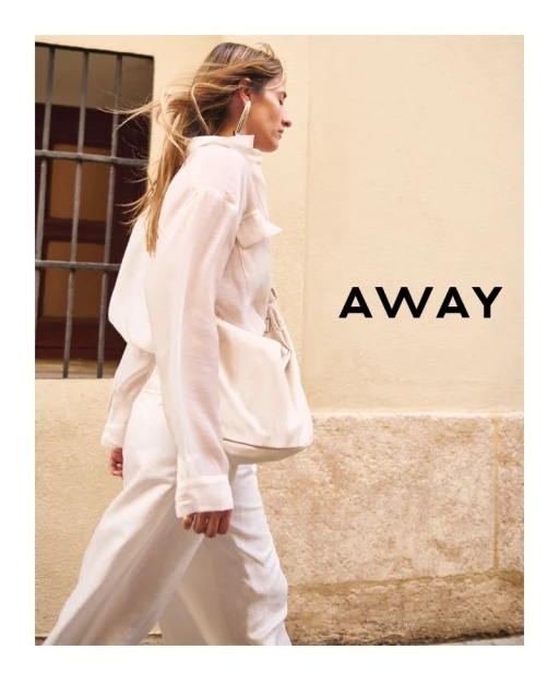 A woman in a white outfit walks on a city street next to a wall. She carries a white bag, and the word 'AWAY' is visible.