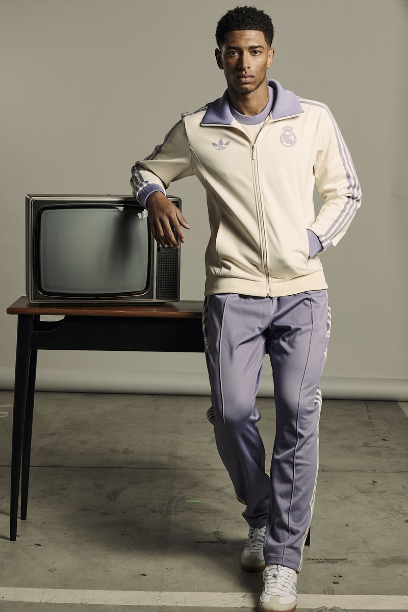 An adult male in a cream and lavender tracksuit poses next to a vintage television set against a light grey background in a studio shot.