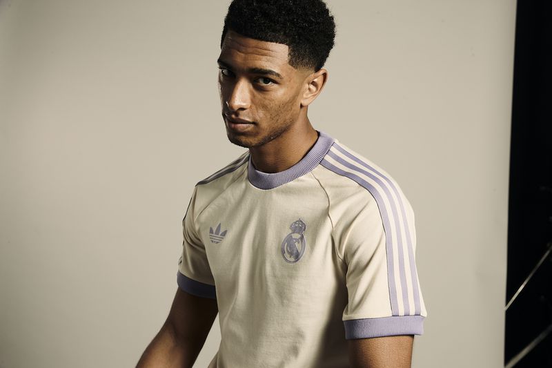 Portrait of a young adult male with dark hair, wearing a beige t-shirt with lilac stripes and the Real Madrid logo against a gray background.