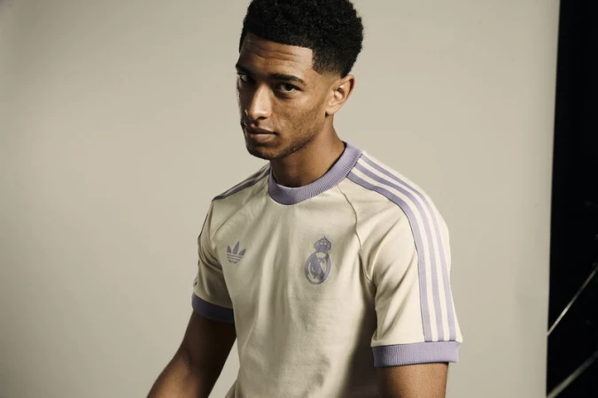 Portrait of a young adult male with dark hair, wearing a beige t-shirt with lilac stripes and the Real Madrid logo against a gray background.