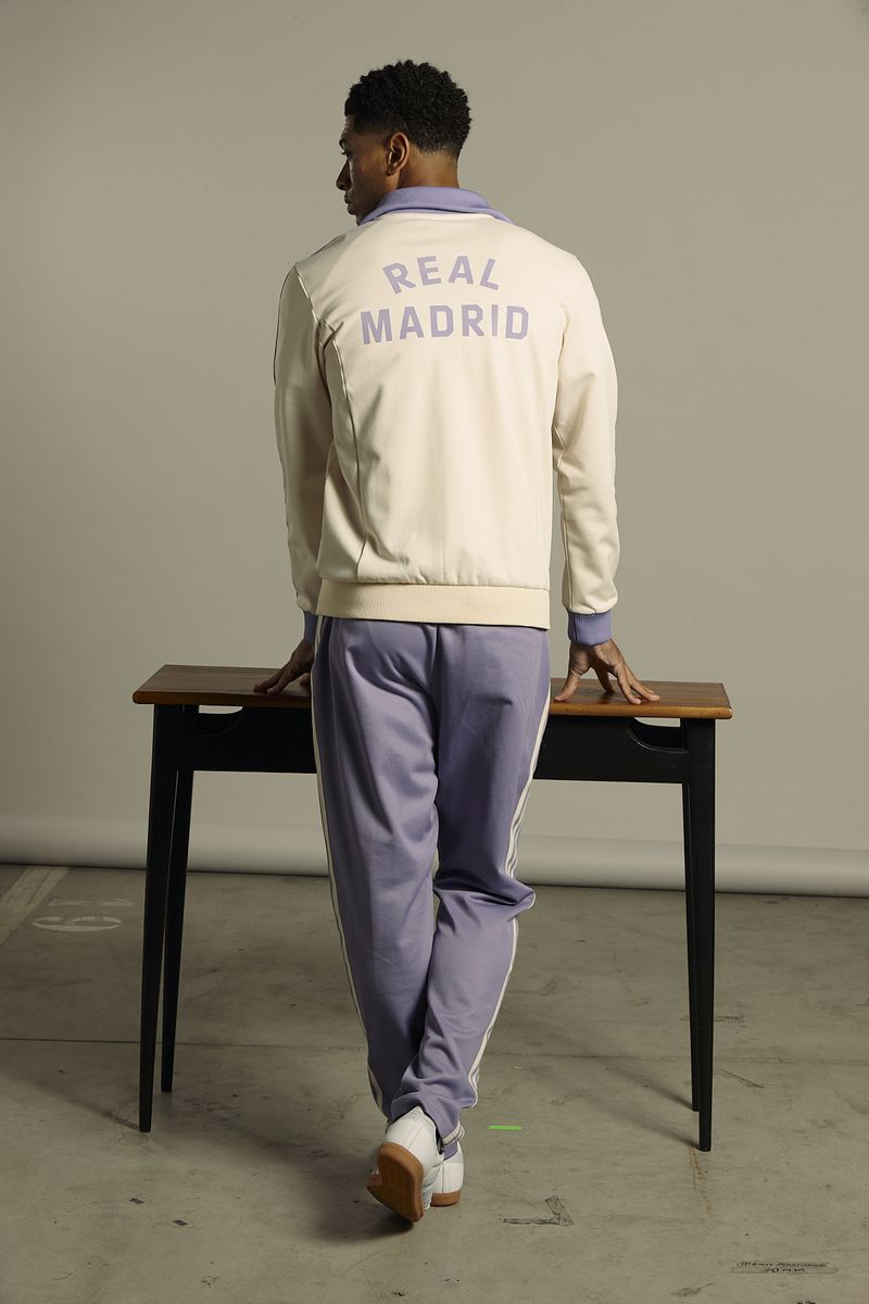 Rear view of a man in a tracksuit with 'Real Madrid' printed on the back, posing next to a desk in a neutral studio setting.