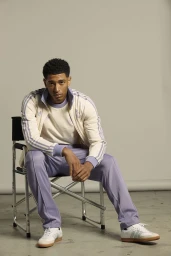 Jude Bellingham seated on a director chair wearing an Adidas tracksuit against a neutral studio background