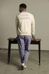 Jude Bellingham facing away, wearing an Adidas Real Madrid cream and purple tracksuit standing at a dark table against a grey seamless backdrop