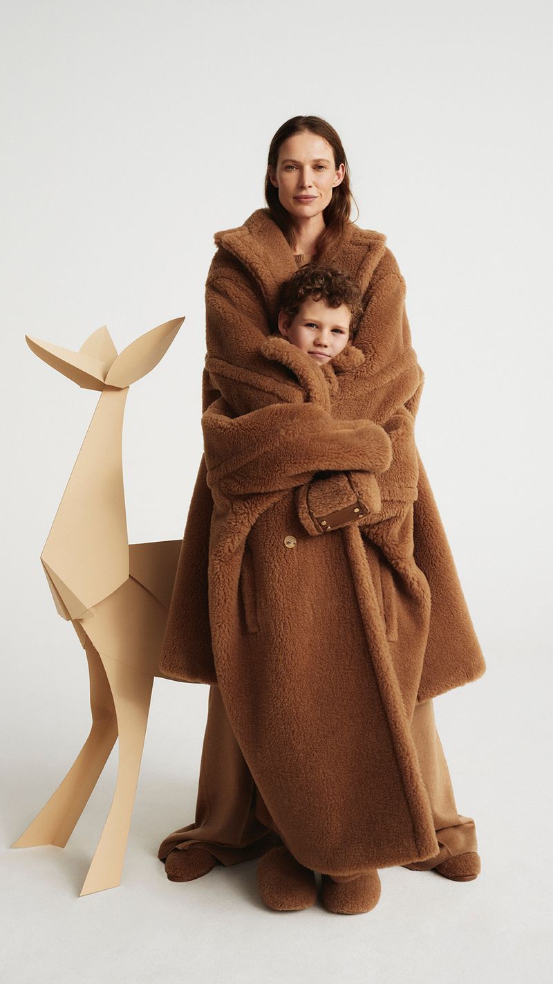 Woman and child in matching brown teddy bear coats embrace beside a tan paper deer cutout against a stark white backdrop.