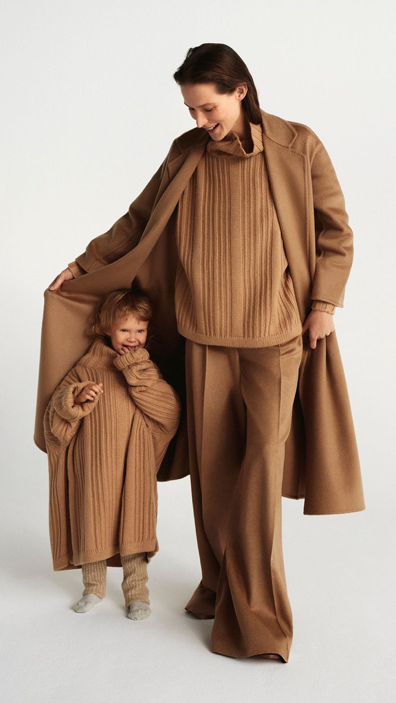 Adult woman and a toddler in matching tan knit outfits posing in a photo studio against a white backdrop, with a long tan coat.