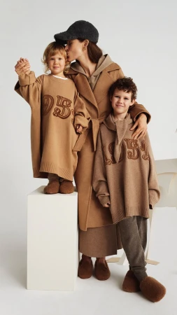 Mother with two children in matching brown knitwear outfits and fuzzy slippers posing on white block in a bright, minimalist photo studio.