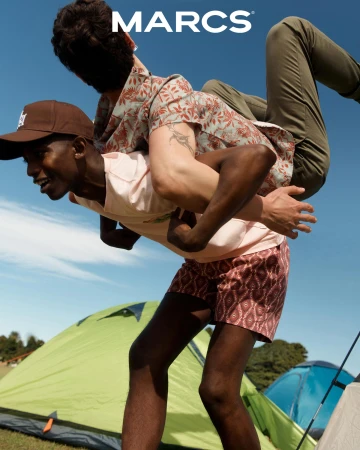 Two young adult men outdoors in a playful piggyback ride, showcasing patterned shorts and a hat against a clear blue sky and camping tents.
