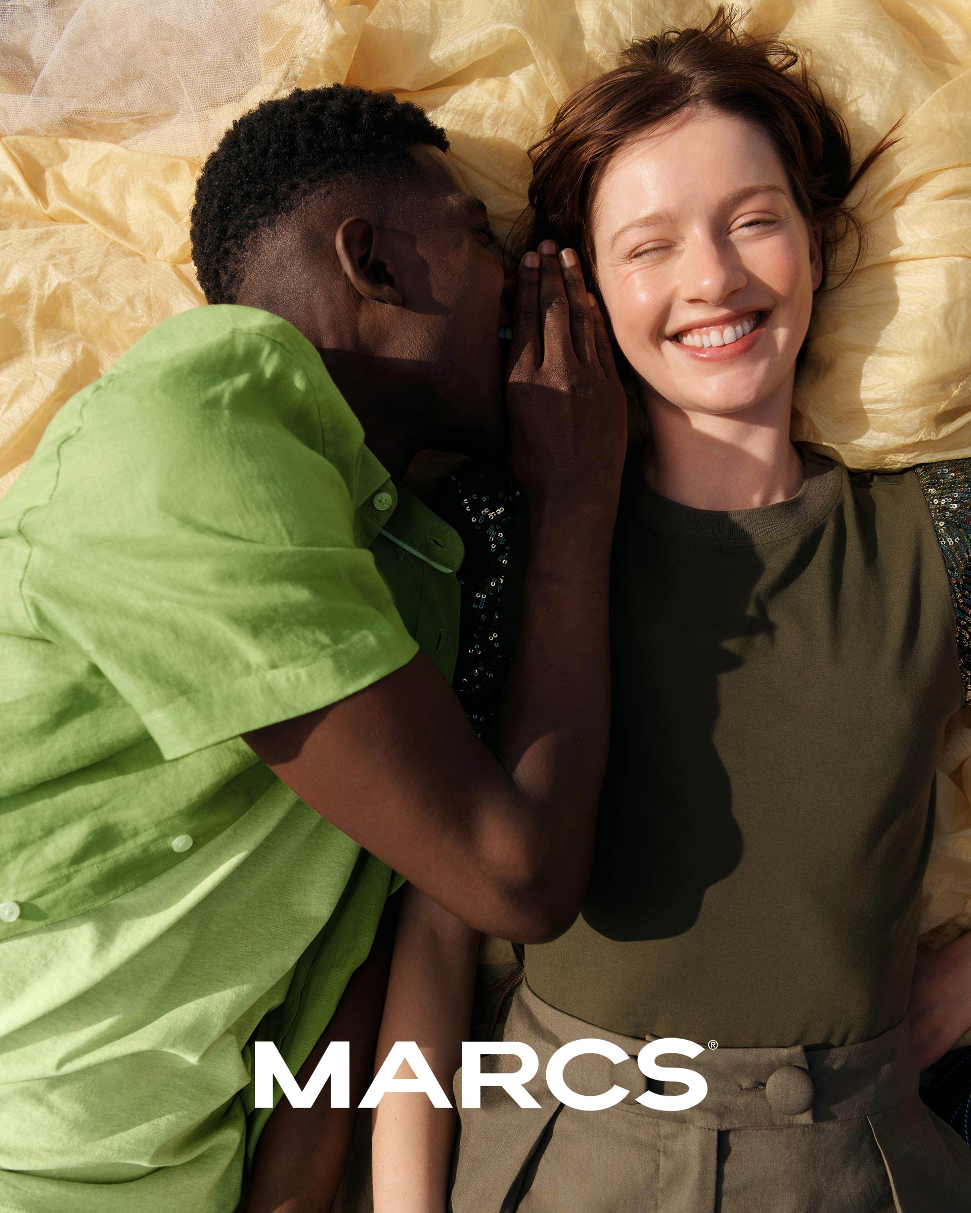 An African-American man whispers to a Caucasian woman with red hair smiling in a fashion shot, posing on a yellow and floral patterned blanket.
