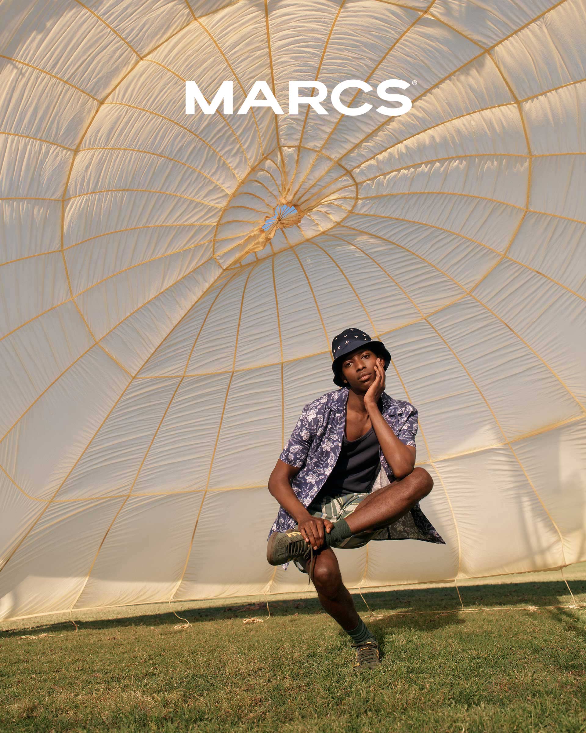 Young adult poses beneath a large, patterned dome structure in casual summer fashion, sitting on the grass in an outdoor setting.