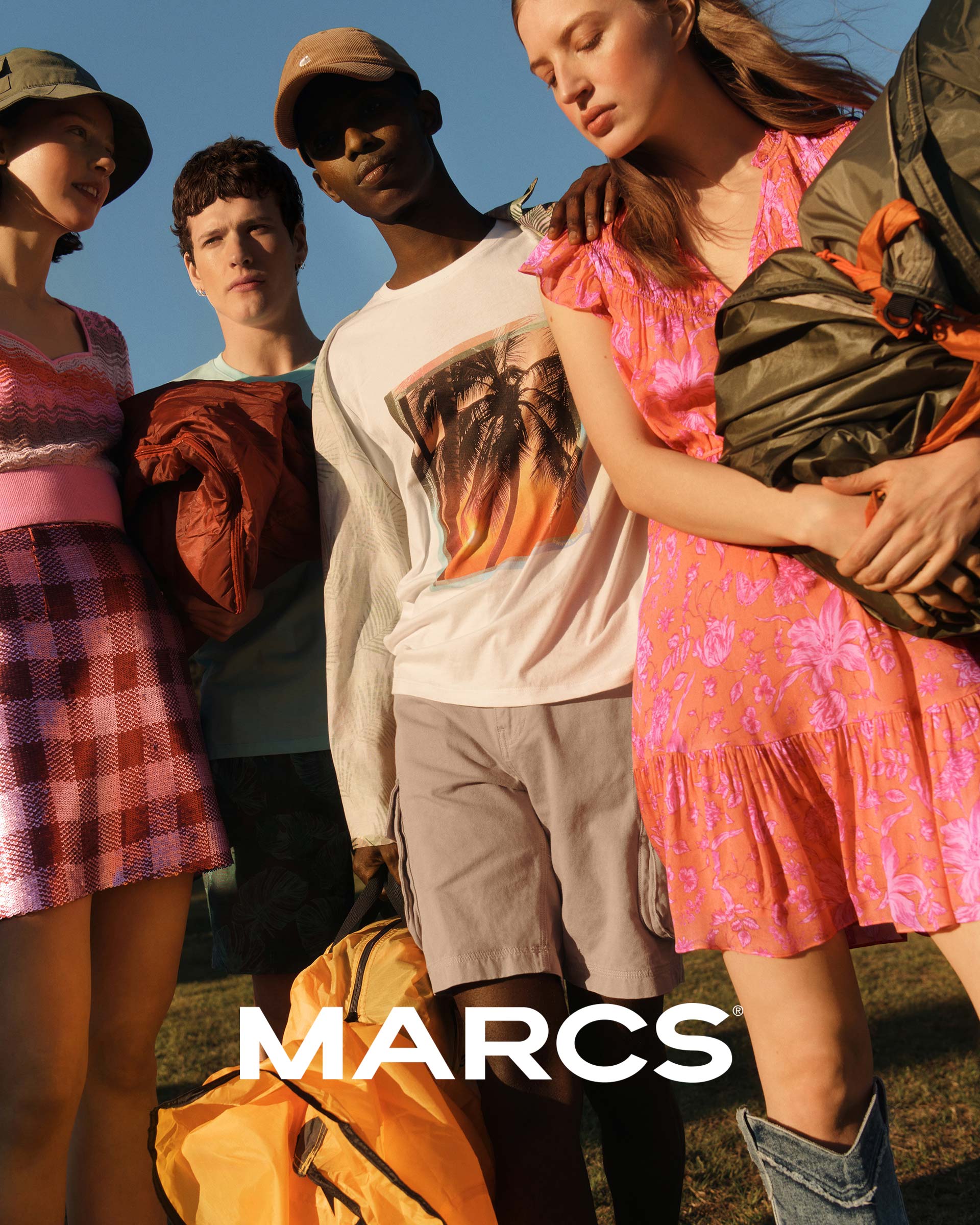 Group of young adults in fashionable summer clothes, including printed dresses and shorts, gathered outdoors with camping gear, posing casually.