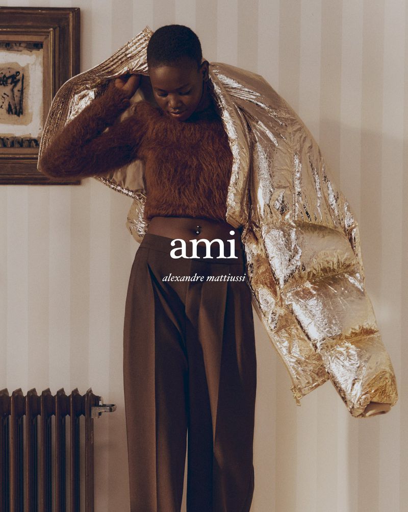 Fashion shot of a Black female model indoors. She is wearing a brown top, brown pants, and a gold puffer jacket. 