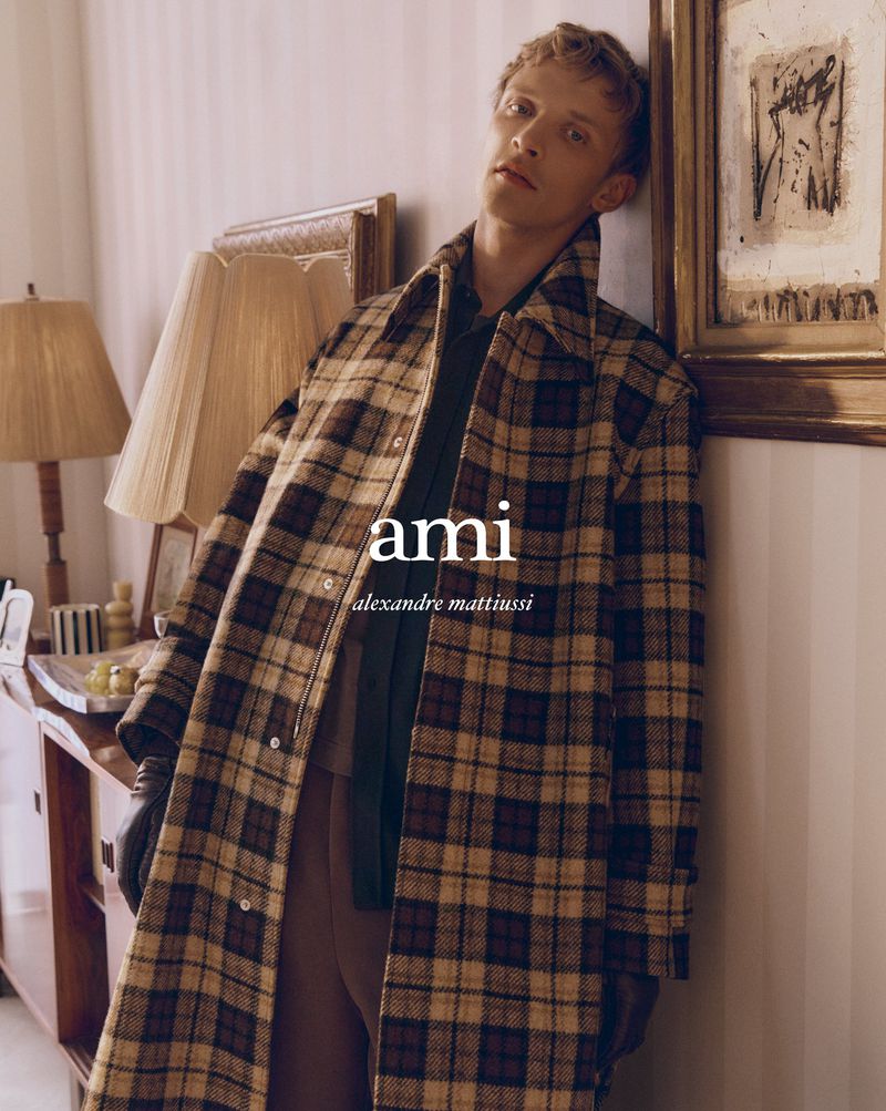 Man in a brown and beige plaid coat leans against a wall next to a wooden desk, lamp, and framed art in an indoor setting.