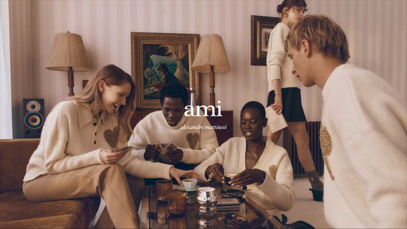 Group of stylish young adults in cream-colored sweaters with heart details gather in a cozy living room, playing cards and drinking tea.
