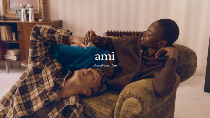 Two people lounge in a warmly lit room, a Black woman in brown hoodie rests against an olive chair, while a white man in plaid lies below.