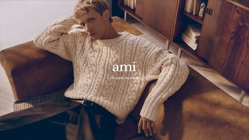 A blond male model poses in an off-white cable-knit sweater and brown pants, seated on a brown couch with a wood cabinet behind him.