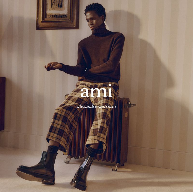 An adult African male model is seated indoors wearing a brown turtleneck, plaid pants, and black boots in a fashion editorial shoot.