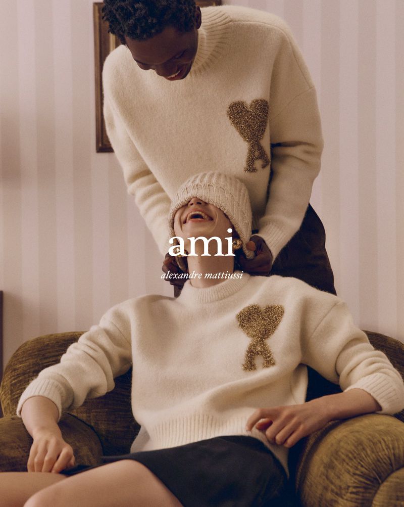 Two people wearing Ami Paris sweaters pose indoors; one sits on an olive-green chair and the other stands behind and smiles downward.