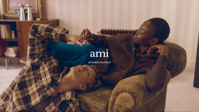 Two models relaxing together indoors, one lying on an armchair wearing a brown hoodie and another reclining on the floor in a plaid shirt