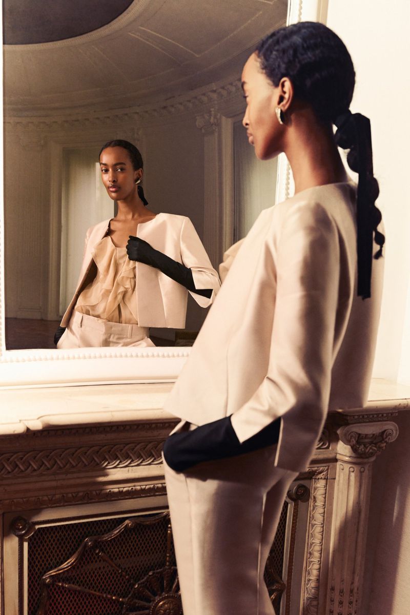 Elegant African-American woman in light peach suit with black gloves gazes at reflection in mirror with ornate carved wooden frame and details.