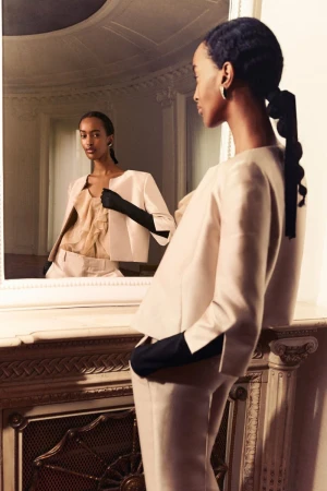 Elegant African-American woman in light peach suit with black gloves gazes at reflection in mirror with ornate carved wooden frame and details.